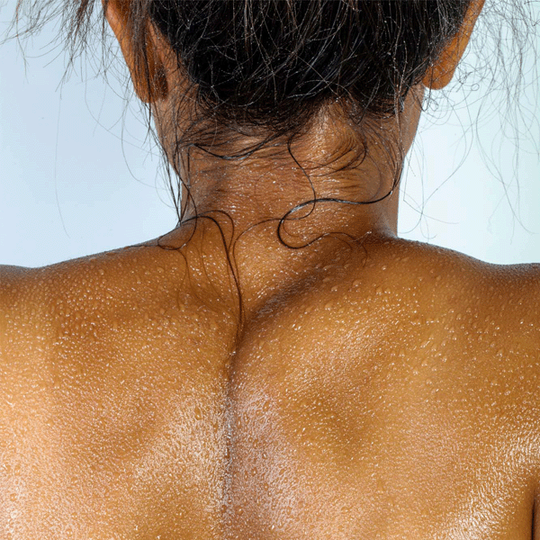 Close-up view of a person's back, glistening with water droplets. The hair is pulled back, revealing skin with a smooth, shiny texture. The background is softly lit, enhancing the wet appearance.